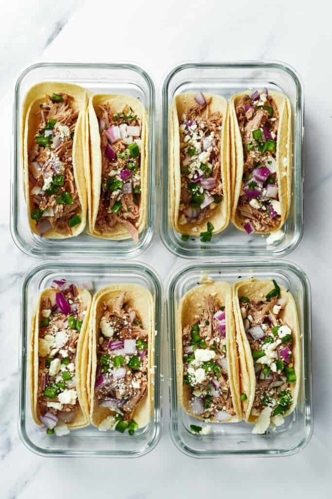 Four meal prep containers, each containing two tacos on corn tortillas with Instant Pot carnitas, jalapeno, red onion, and cojita cheese.
