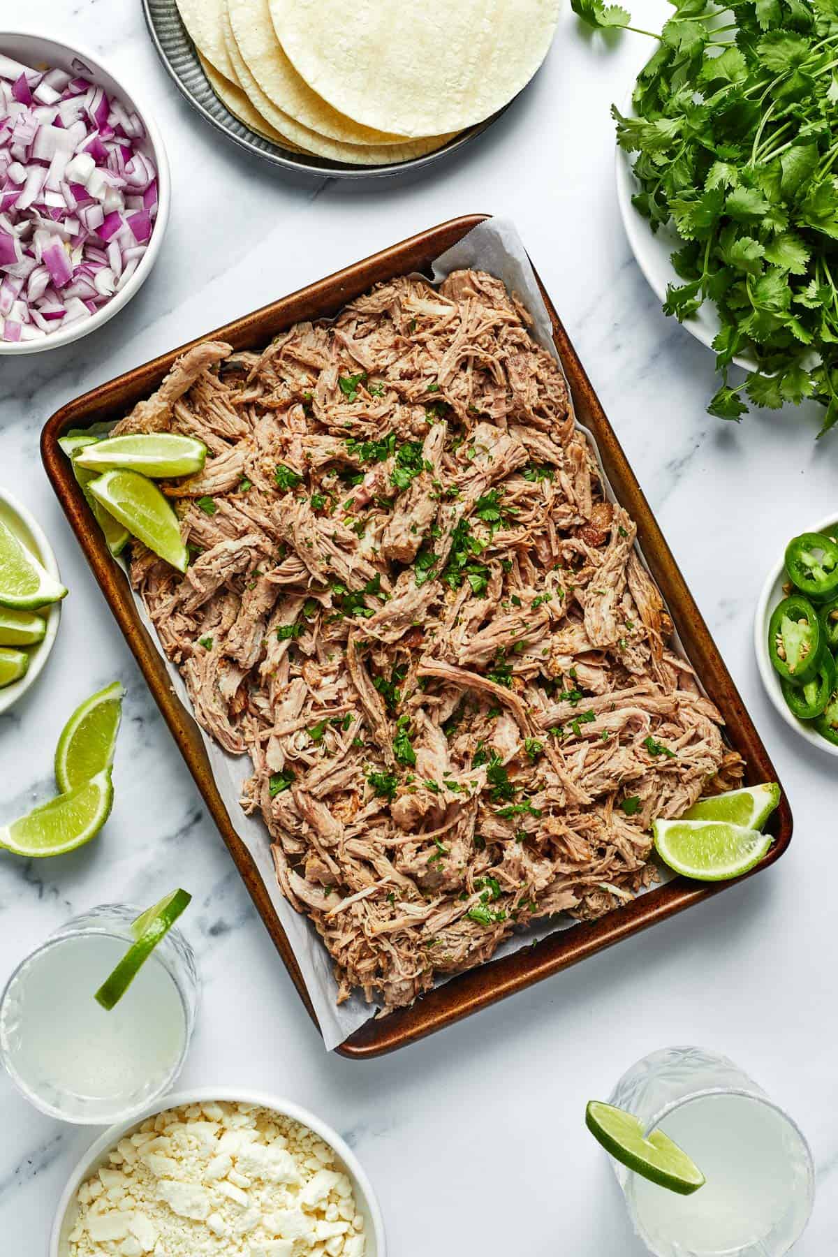 A sheet pan with shredded Instant Pot Carnitas meat surrounded by tortillas, cilantro, limes, red onions, and cojita cheese.
