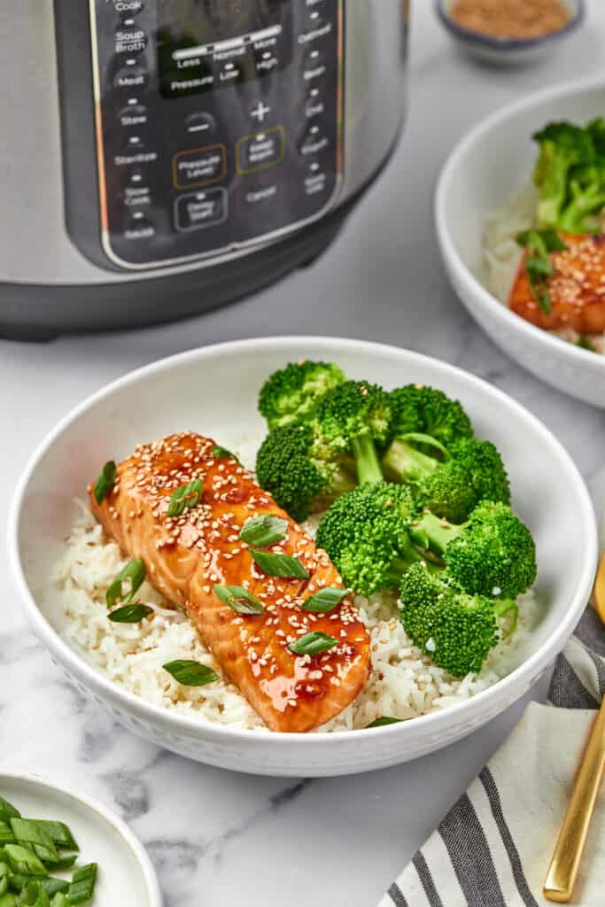 An Instant Pot in the background with a bowl of rice topped with broccoli and teriyaki salmon in the foreground.